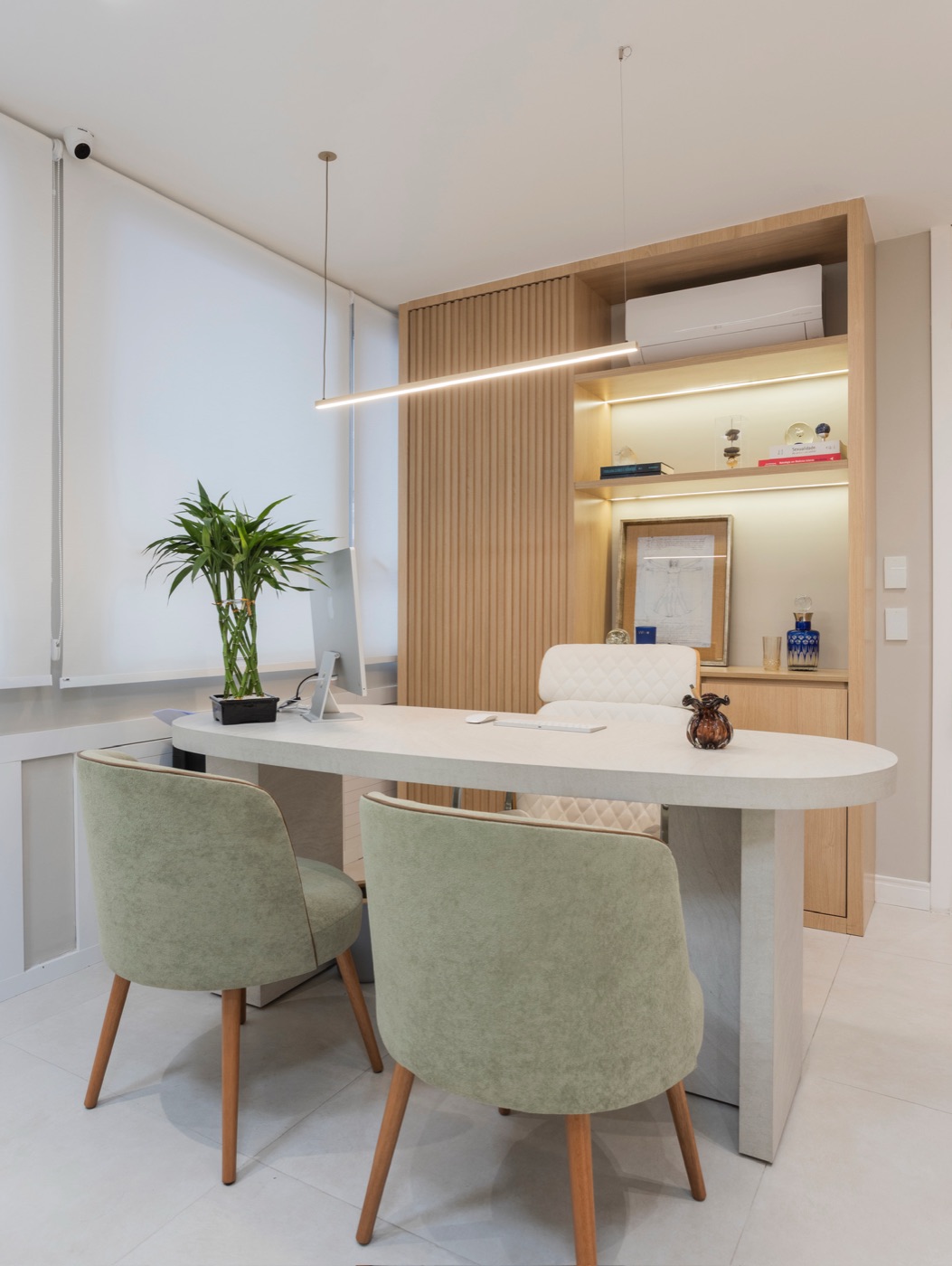 Consultório com mesa oval, cadeiras verdes e estante iluminada
